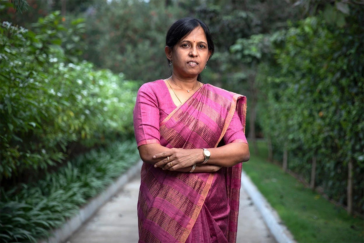 Picture of Kalpana Kalahasti in a pink-colored Indian saree. She is standing on the track of a garden.