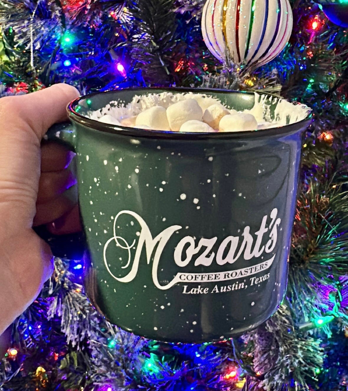 A photo of Shohreh's hand holding up a green mug filled with marhsmallows, marshmallow fluff, and hot chocolate in front of a brightly lit Christmas tree