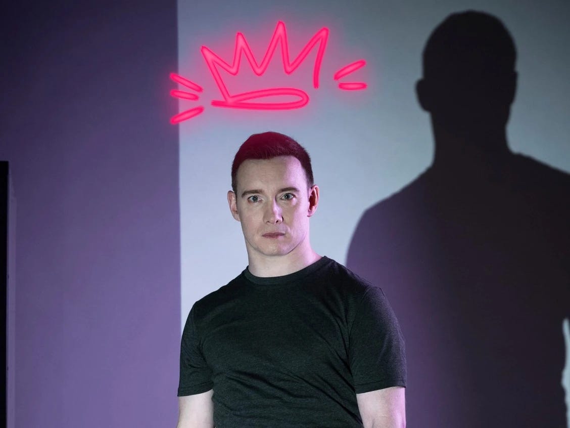 A white man looks squarely at the camera. A neon line drawing of a crown float above his head.