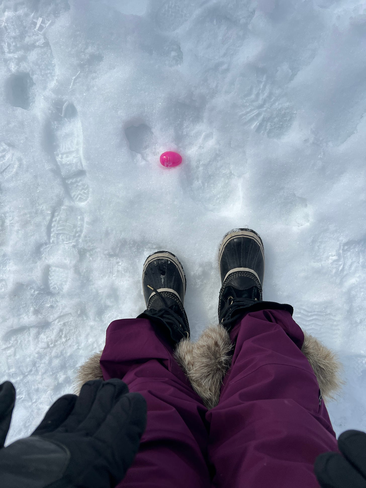Molly standing in the snow in heavy boots, snow pants and gloves, looking down at a pink plastic Easter egg in the snow.