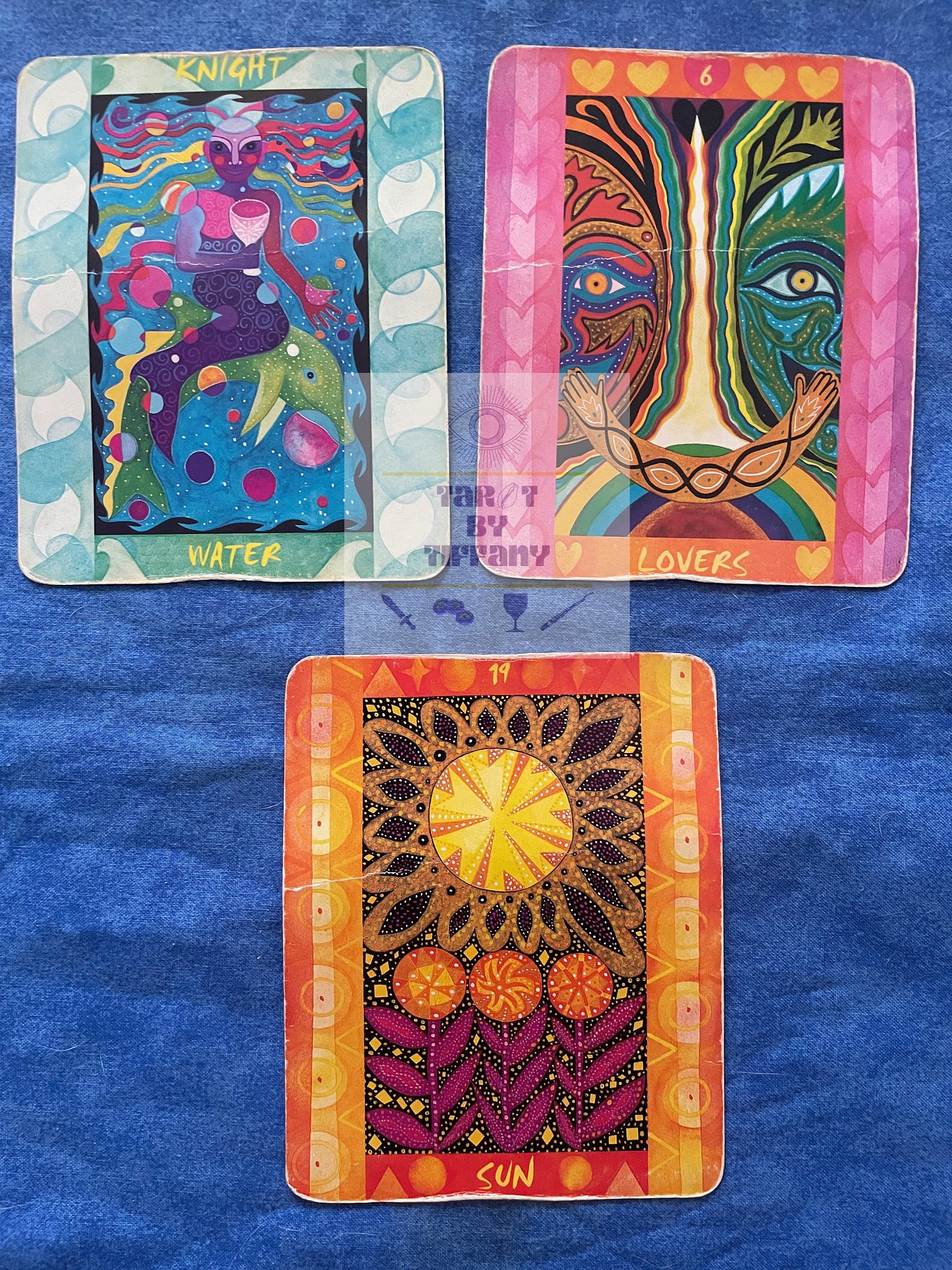 3-card reading with the Tarot of the Four Elements. The cards are laid on a cobalt blue cloth background. Top two cards (left to right): Knight of Water, The Lovers. Bottom card: The Sun.