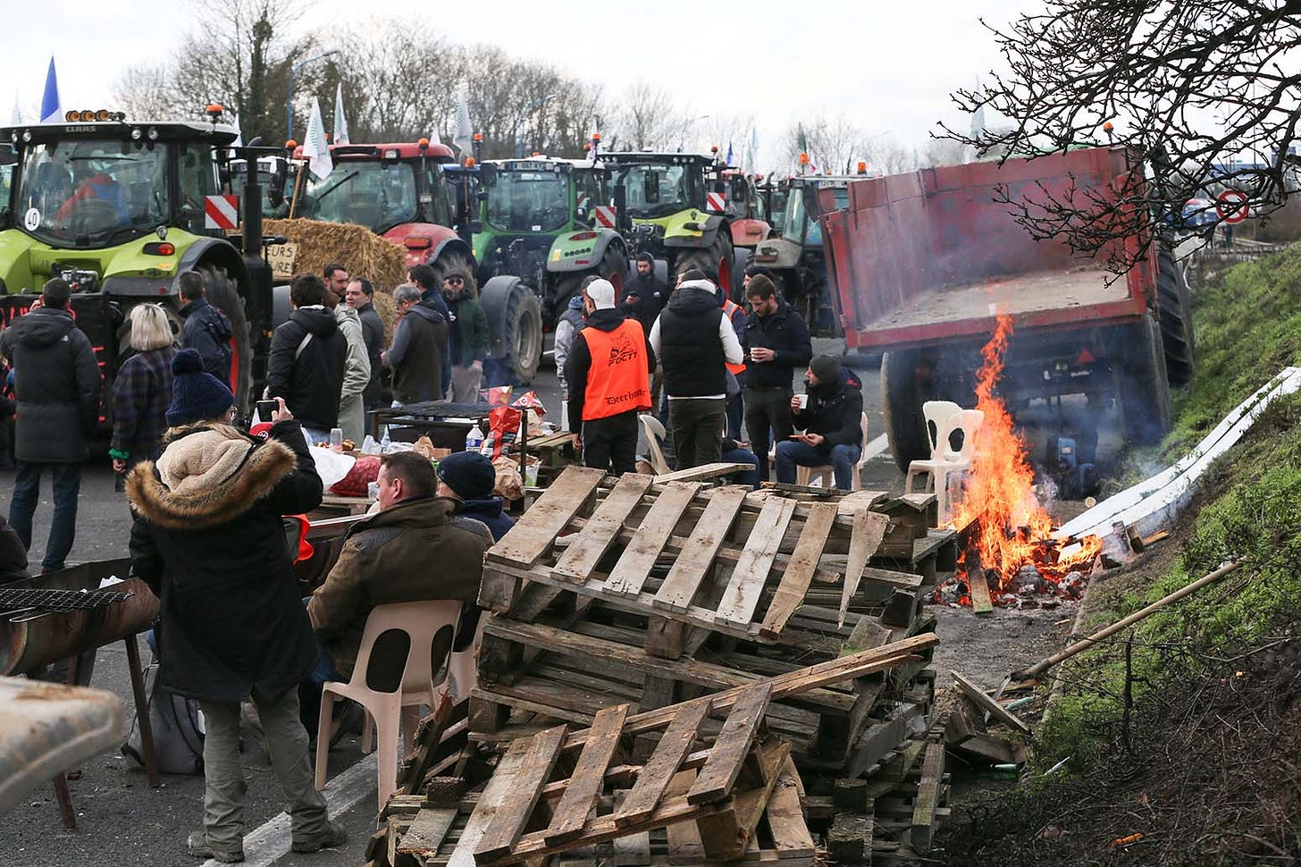 Tractors, bales of hay, wooden shipping pallettes being burned in an open fire, people standing around or sitting on folding chairs, all on a road.