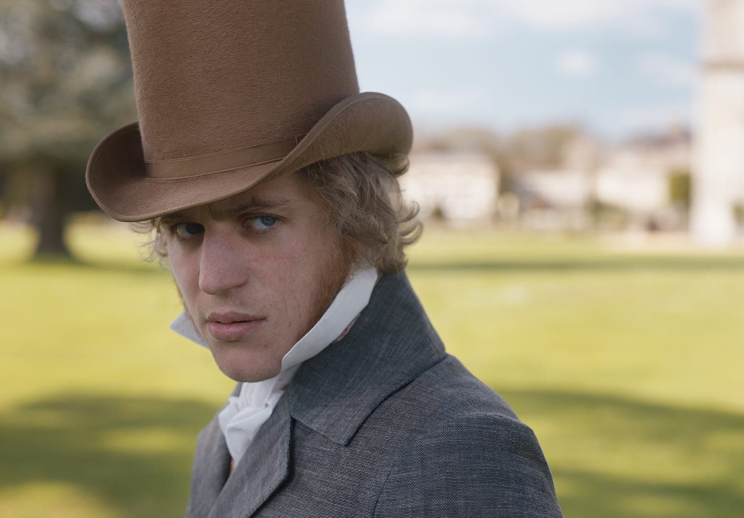 Mr. George Knightley (played by Johnny Flynn in Autumn de Wilde’s film ‘EMMA.’ wears a top-hat and grey suit and looks on with a look of consternation, amidst a background of English lawn. 