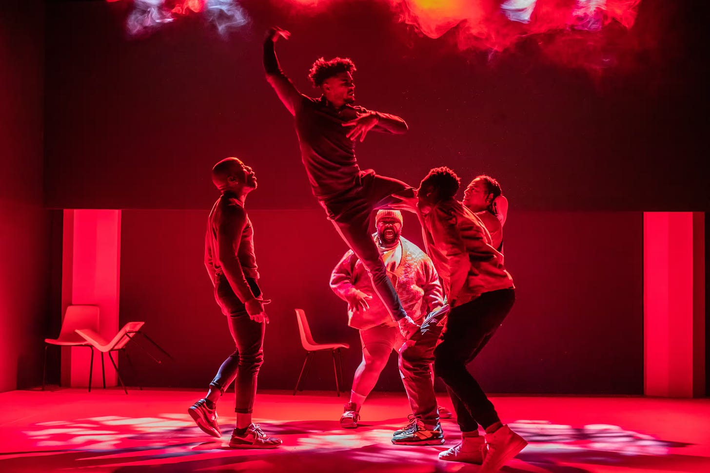 Five Black men actors in a circle, facing each other on a darkened stage bathed in red light. The central actor is mid leap.