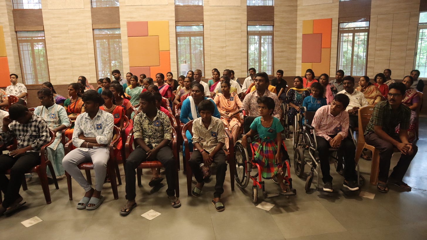 The image shows a group of students and their family members gathered in the Vidya Sagar Auditorium. The students are seated in several rows of red plastic chairs, with some in wheelchairs. They are participating in a celebration called the "Rising Star" event. The atmosphere is filled with pride and joy as the family members, dressed in colorful traditional attire, look on with happy and proud expressions. The walls of the auditorium are decorated with square and rectangular panels in various warm colors, adding to the festive mood. Large windows with wooden blinds allow natural light to fill the space, creating a warm and welcoming environment.