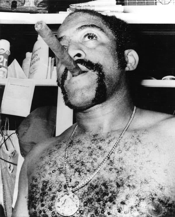 A black and white close-up photo of a shirtless Tiant smoking a big cigar in a locker room. He has a chain with a medallion draped around his neck.