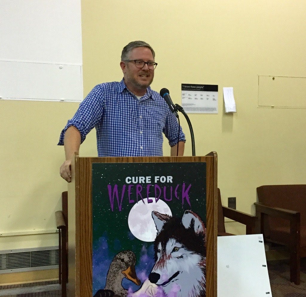 A photo of me from 2016. I'm a 30-something white guy with a beard. I am speaking at a lecturn with a microphone. On the front of the lectern is an oversize image of my second book "cure for wereduck"