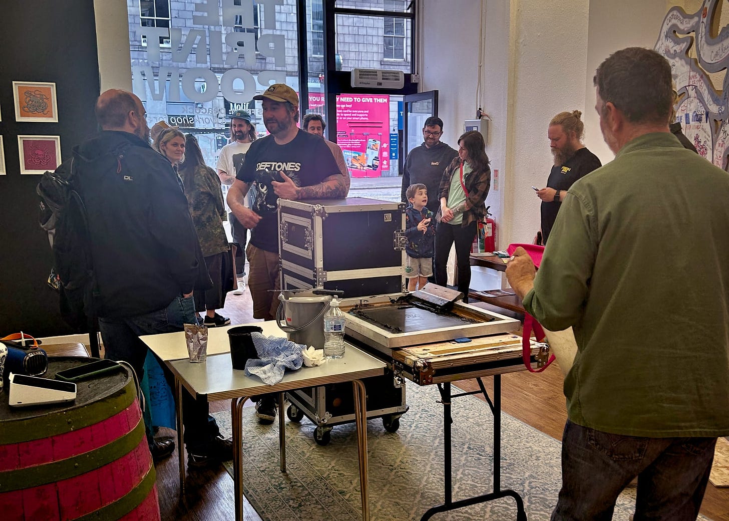 A photo from the opening of Craig Fisher’s 'Popcorn Octopus' show at The Print Room. The room is bustling with visitors of various ages engaged in conversation. In the center, a man wearing a band t-shirt is interacting with attendees near a printmaking station with equipment and ink, suggesting a live demonstration. Artwork adorns the walls, and the venue has an inviting atmosphere with natural light coming in from the large windows.