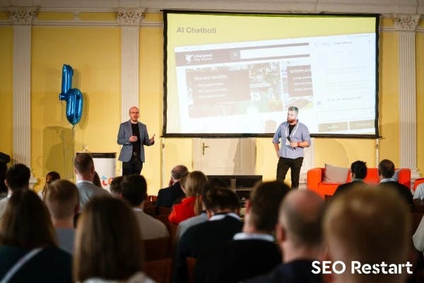 Martin Kopta (vlevo) a Jakub Goldmann na pódiu konference SEO Restart a za nimi slajd s AI Chatbotem na stránkách liverpoolské radnice.