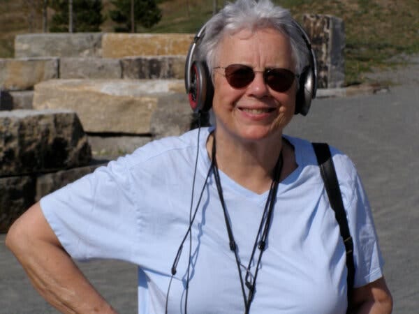 Helen Thorington, an older woman with short white hair, wearing headphones, sunglasses and a light blue T-shirt, standing outdoors.