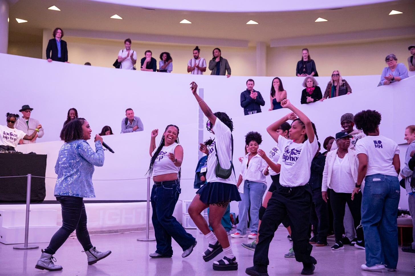 Dancers in Ladies of Hip-Hop t-shirts smile and put their hands in the air, facing an emcee with a mic and surrounded by people looking on and clapping.