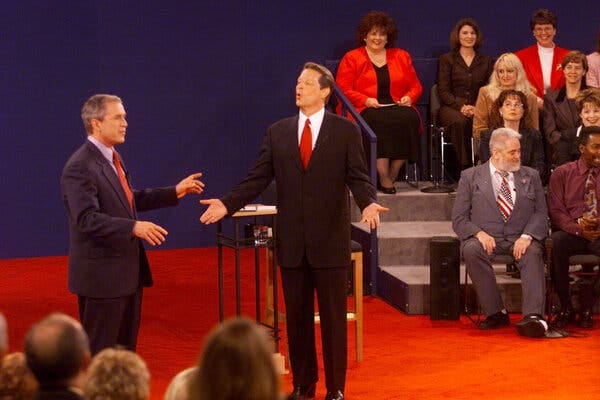 Vice President Al Gore, right, is speaking while standing next to Gov. George W. Bush as an audience seated around them watches.