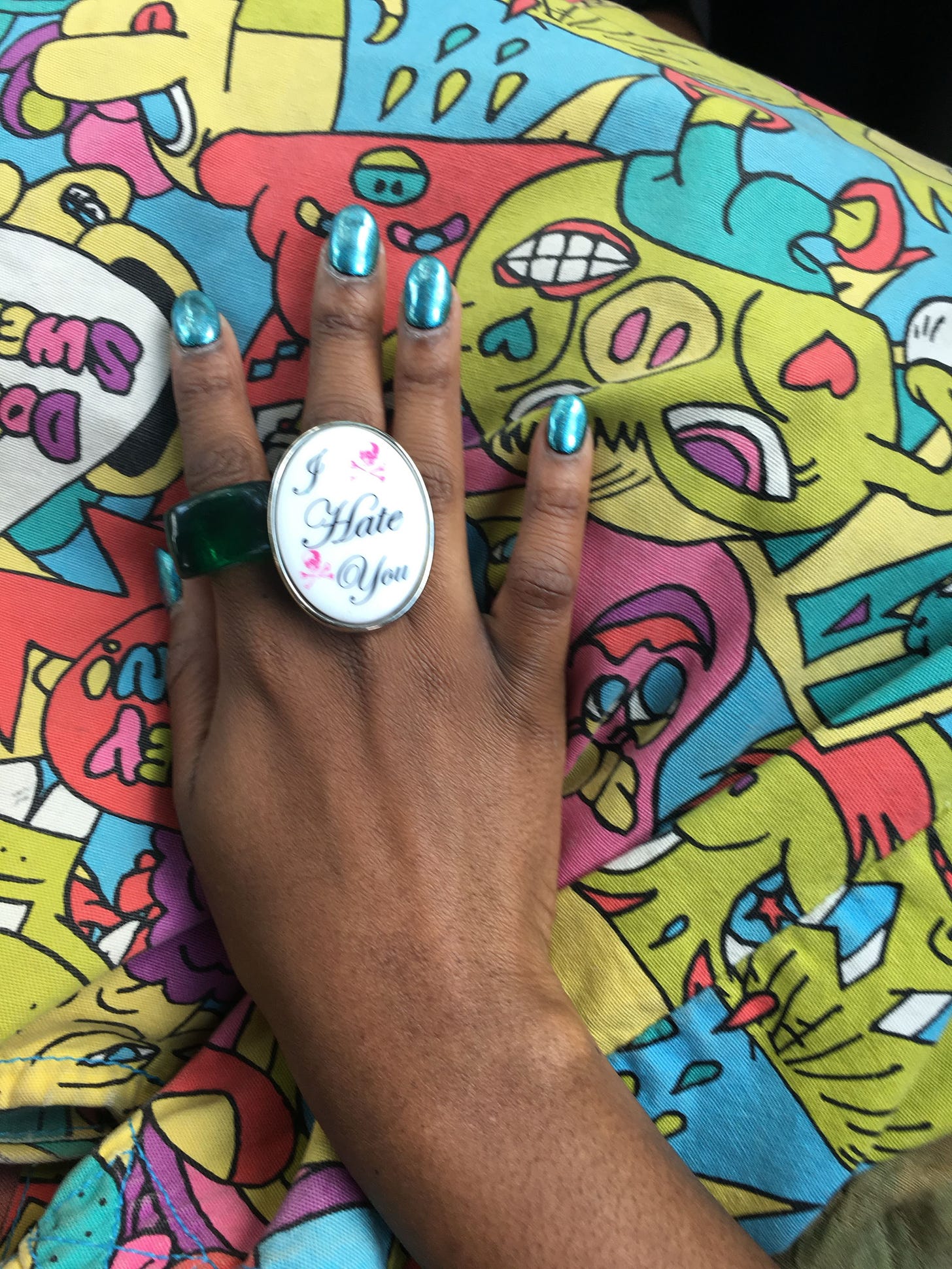 a Black woman’s hand rests on a colorful bag. the person wears a blocky green ring and another large ring that says “I Hate You.”