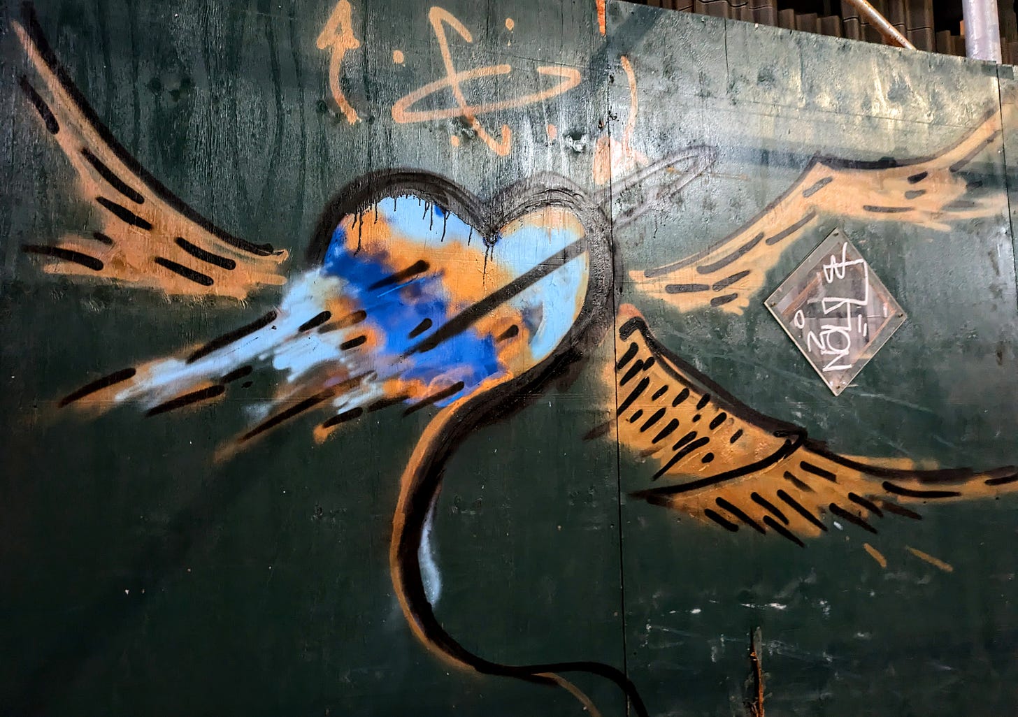 A heart spray-painted on a green wall. The heart has orange wings and blue streaks flowing out of it. 