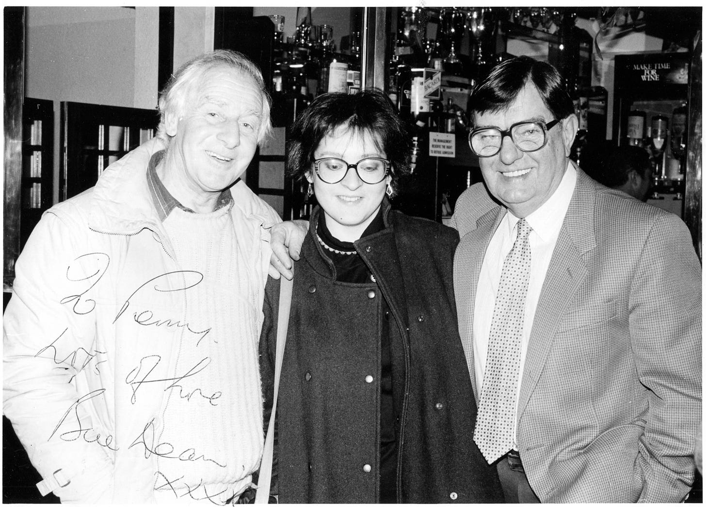 Black and white photo of a young woman, looking shy, standing between two men, looking cheerful. The photo is autographed with the words "To Penny, Lots of love, Bill Dean xxx".