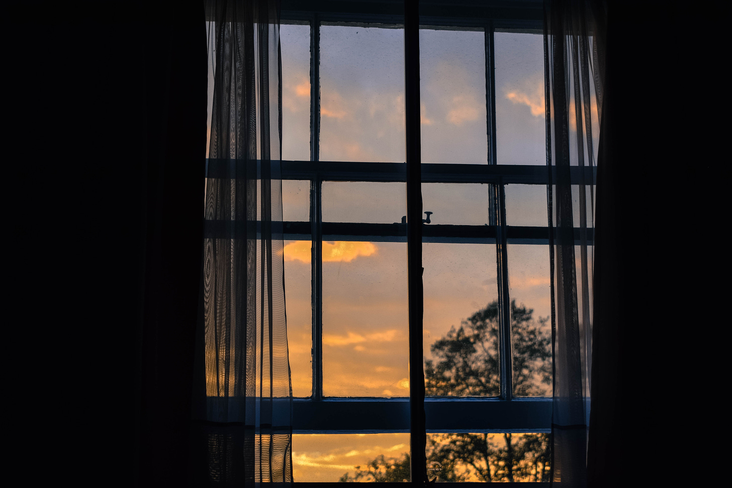 View from within a dark room, looking out an open window on a sunset scene. The sky shades from pale blue to gold.