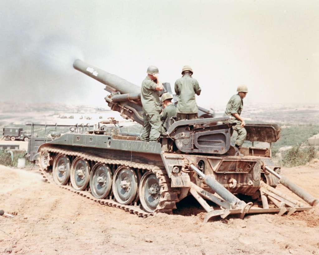 Color photo from March 17, 1968 in Vietnam of a self-propelled heavy artillery gun firing. The self-propelled gun is facing away from the point of view parked on a red dirt foreground firing into the distance. There are four army gun crew onboard the vehicle dressed in combat fatigues and helmets. Color photo from March 17, 1968 in Vietnam of a self-propelled heavy artillery gun firing. The self-propelled gun is facing away from the point of view parked on a red dirt foreground firing into the distance. There are four army gun crew onboard the vehicle dressed in combat fatigues and helmets.