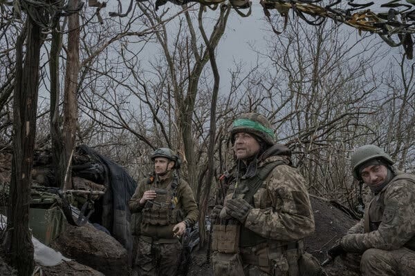 Three Ukrainian soldiers stand together surrounded by trees. 