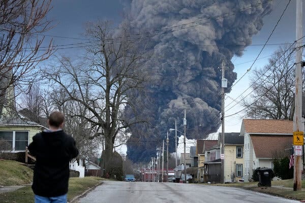 A man in a black jacket looks down a residential street toward an immense column of billowing gray-black smoke in the distance.