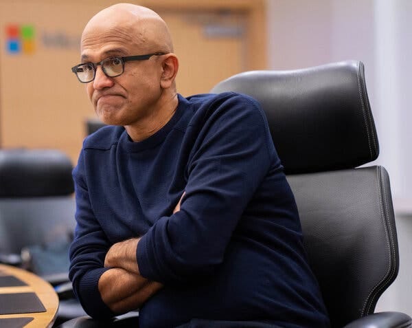 Satya Nadella, wearing glasses and a dark shirt, folds his arms as he sits in an office chair.