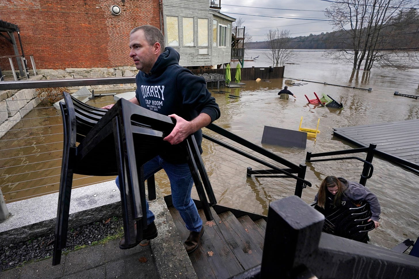 Joe Stanhope and Tori Grasse carry furniture from the flooded outdoor patio of the Quarry Tap Room, Tuesday, Dec. 19, 2023, in Hallowell, Maine. Nathan Stanhope, rear, wades through the flood water to retrieve more items. Waters continue to rise in the Kennebec River following Monday's severe storm. (AP Photo/Robert F. Bukaty)