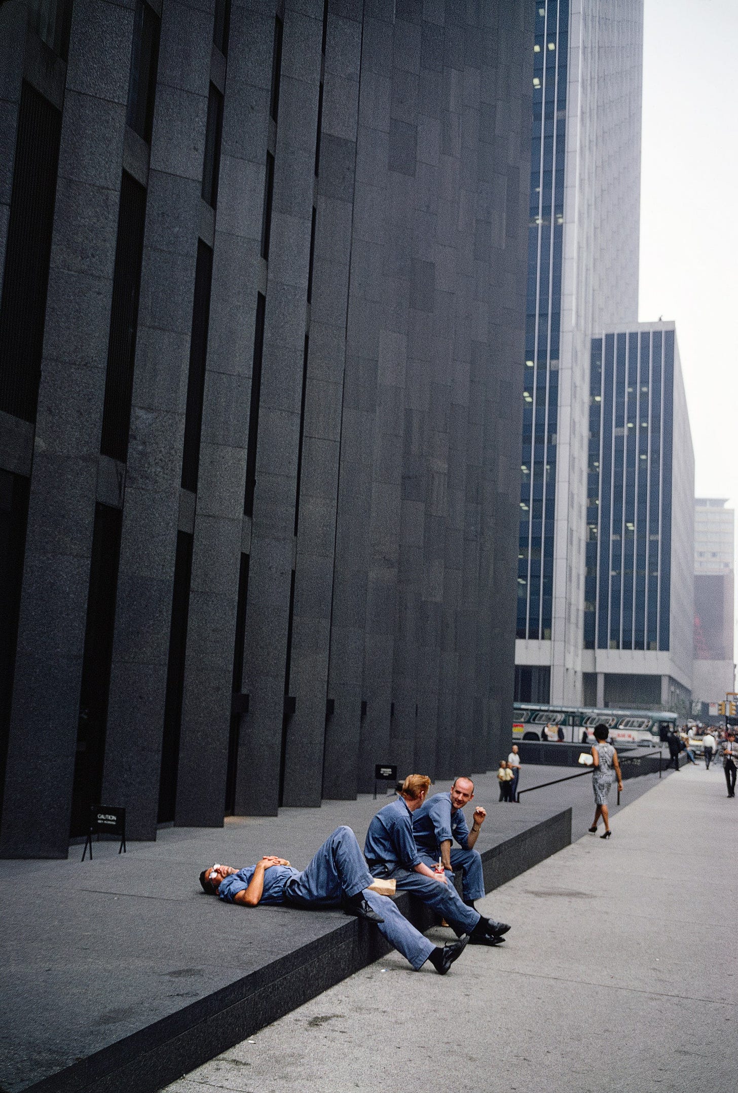 Three people in gray work suits sitting on steps near a skyscraper.
