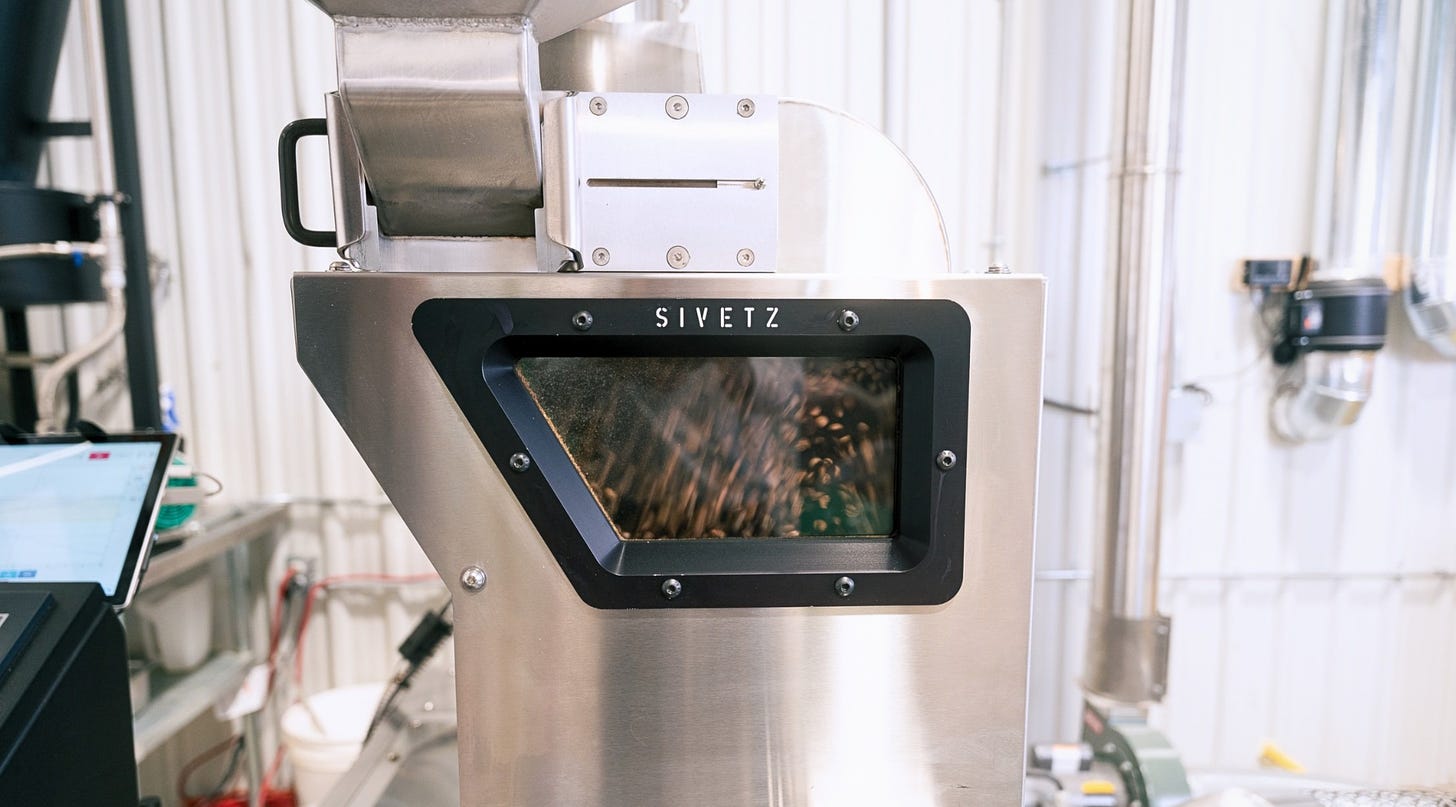 A close-up on the window of a convective air roaster. Coffee beans are bouncing around behind the glass which is framed in black and part of a larger stainless steel machine.