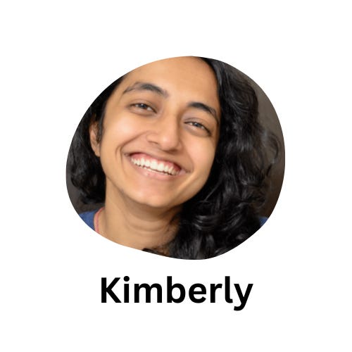 Kim, a brown-skinned nonbinary person, is standing against a blurry grey wall. They have long curly hair that falls past their left shoulder, and are wearing a blue t-shirt. They are smiling at the camera.