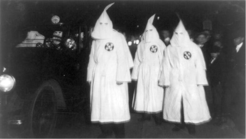 Three men dressed in white robes and white shrouds walk on the street beside a car which is being driven by other men Klan members. Civilians stand beside the Klansmen.