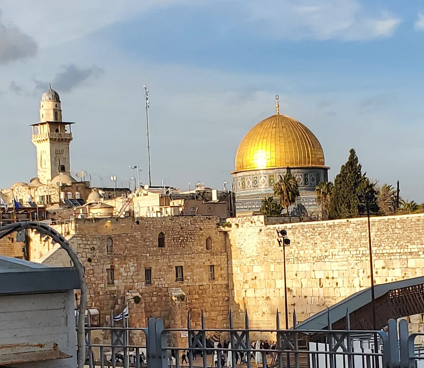 A golden dome and a minaret rise above Jerusalem against a blue sky. 