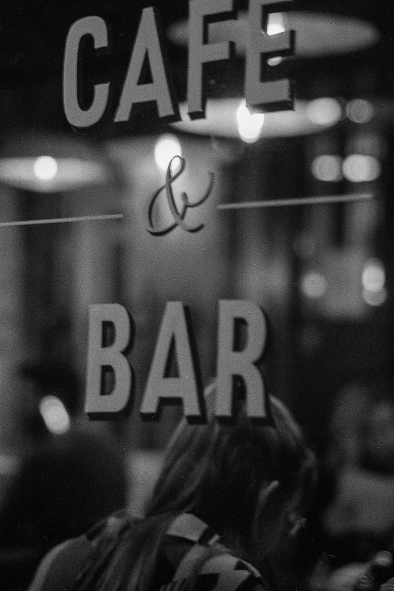 Black and white photo of a woman's back seen through a window with stencil writing saying CAFE & BAR