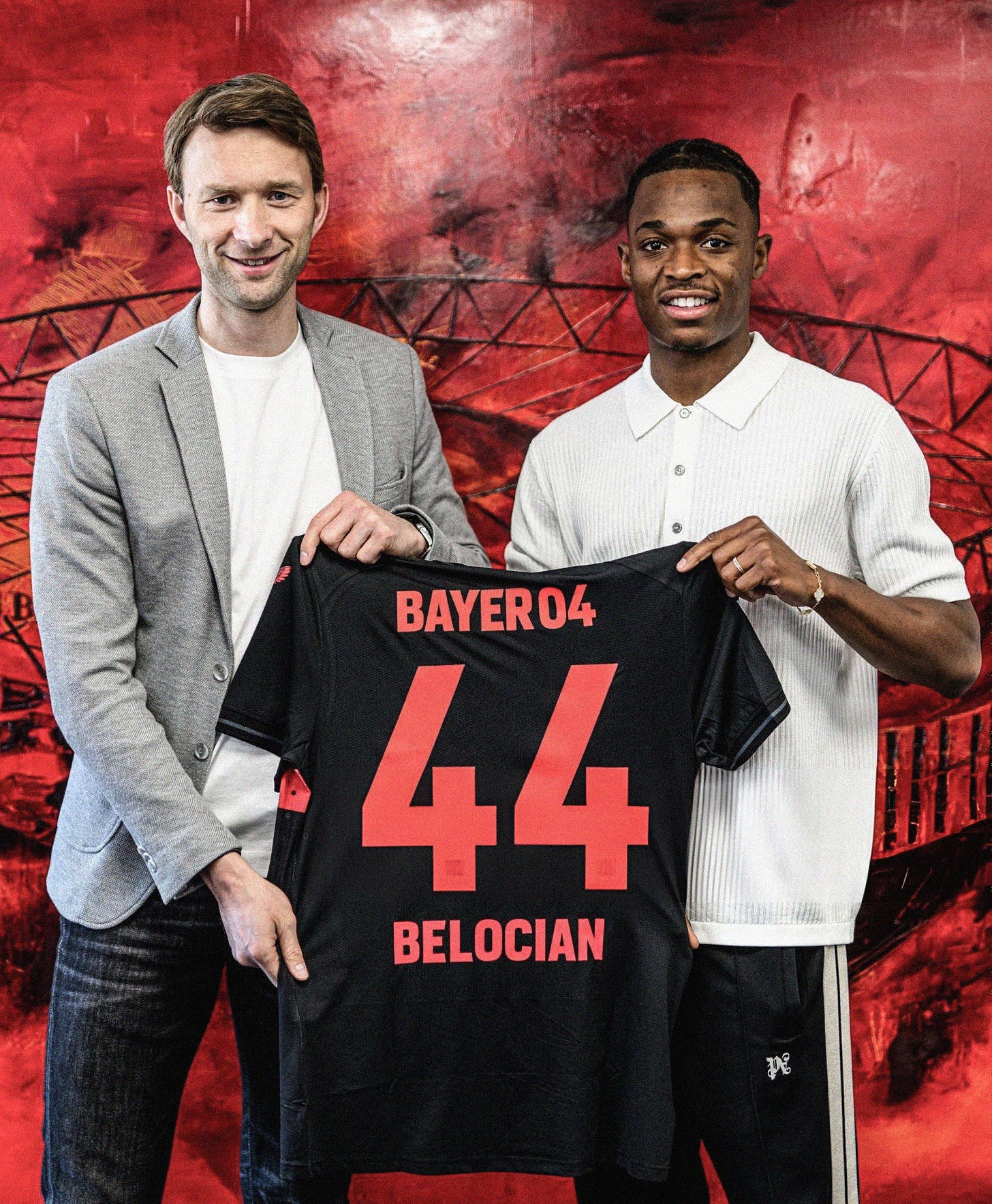 A photo of Jeanuël Belocian holding a black Bayer Leverkusen shirt (with '44 BELOCIAN' in red print on the back) alongside Simon Rolfes. They're stood in front of a wall with a red mural of the Bayer Leverkusen stadium on the back of it.