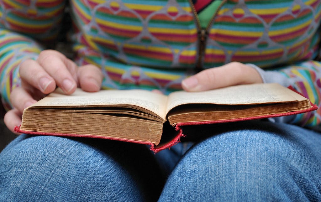 Person wearing jeans and a colorful sweater holds an open book on their lap. ".read" by .brioso. is licensed under CC BY-NC-SA 2.0.