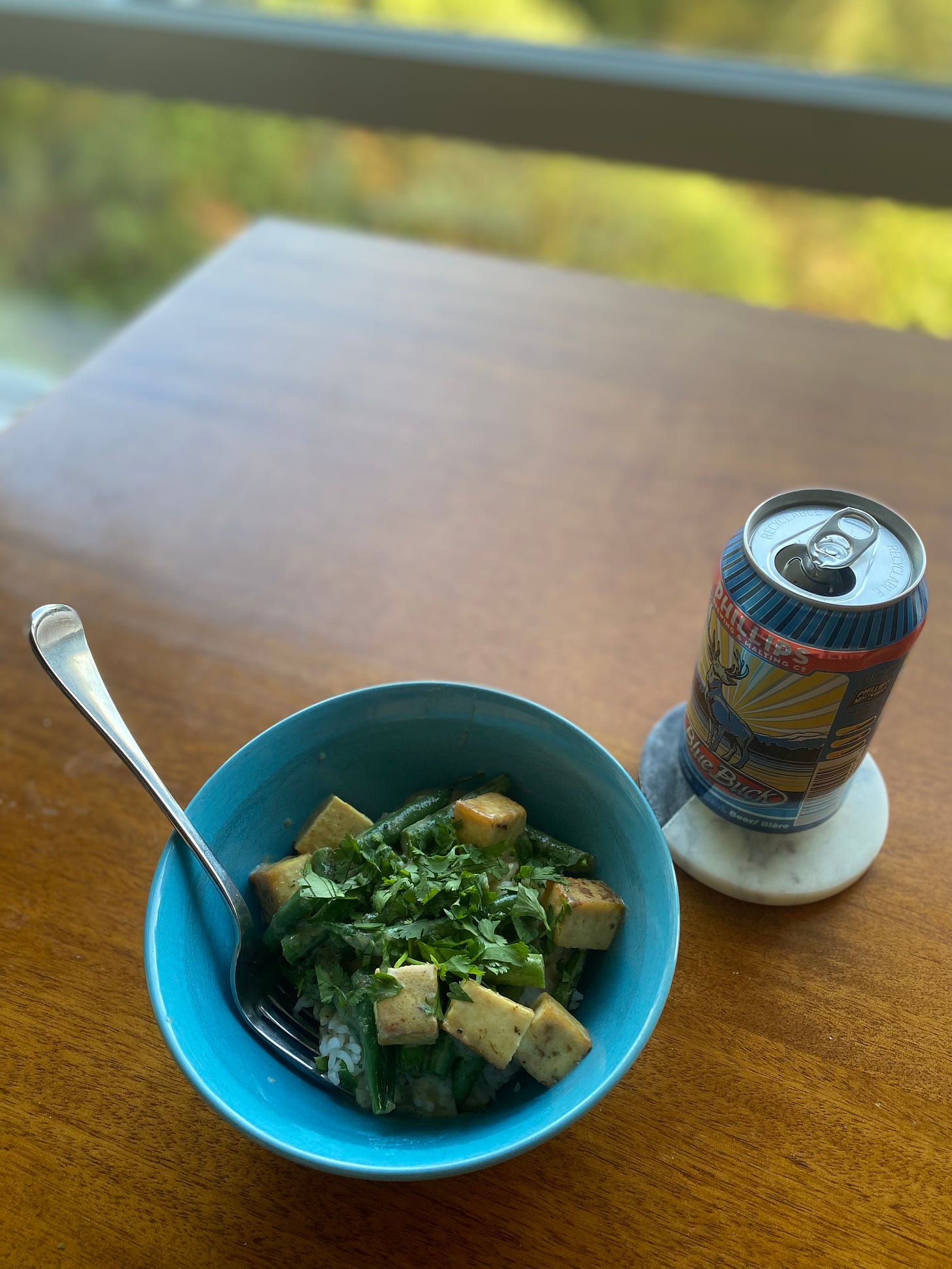 A blue bowl with the curry described above, over jasmine rice. Crispy cubes of tofu and pieces of green beans are visible beneath plenty of chopped cilantro. On a coaster beside the bowl is a can of Phillips Blue Buck pale ale
