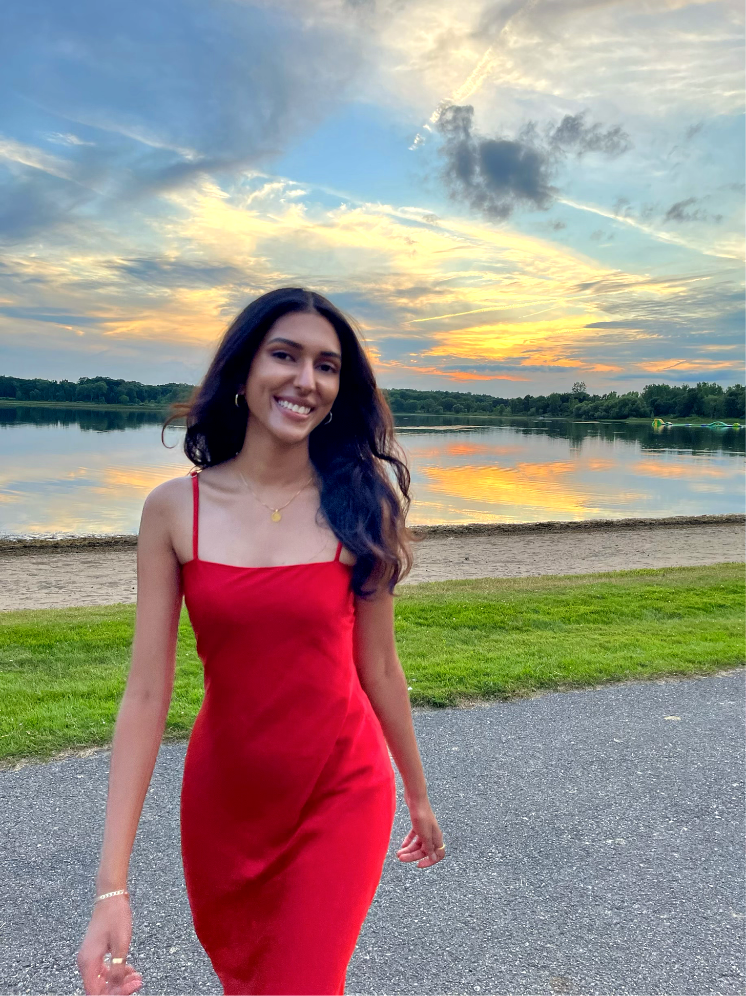 Abi in a red dress walking towards the camera smiling in front of a sunset, water, and some grass.