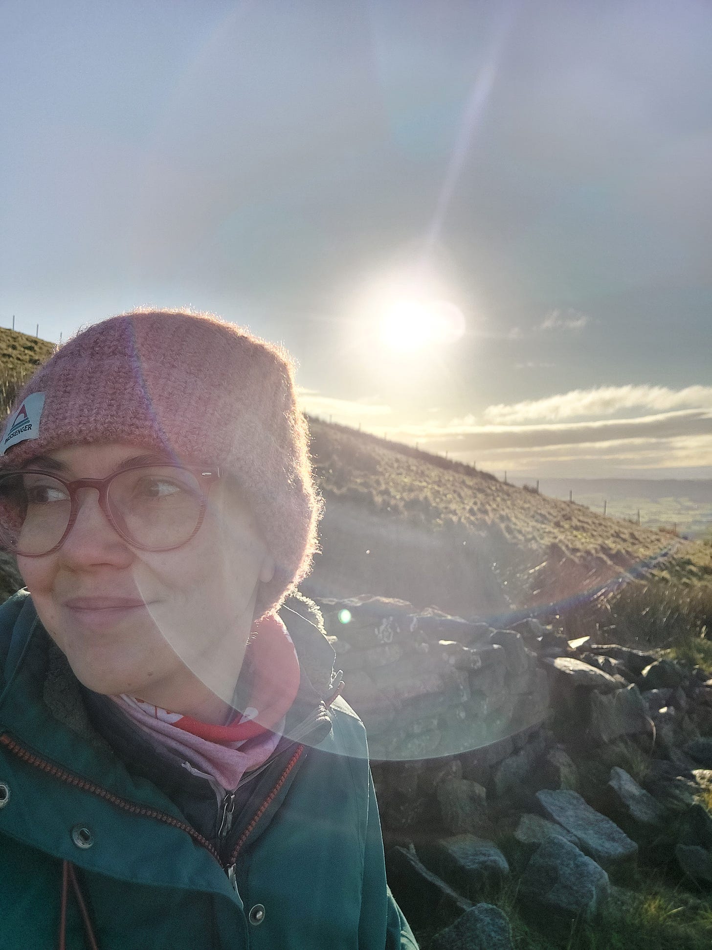 A selfie of me with the dawn sun shining in the background. I've walked high up Parlick hill to take in the view. I'm wearing a pink hat as it was chilly up there