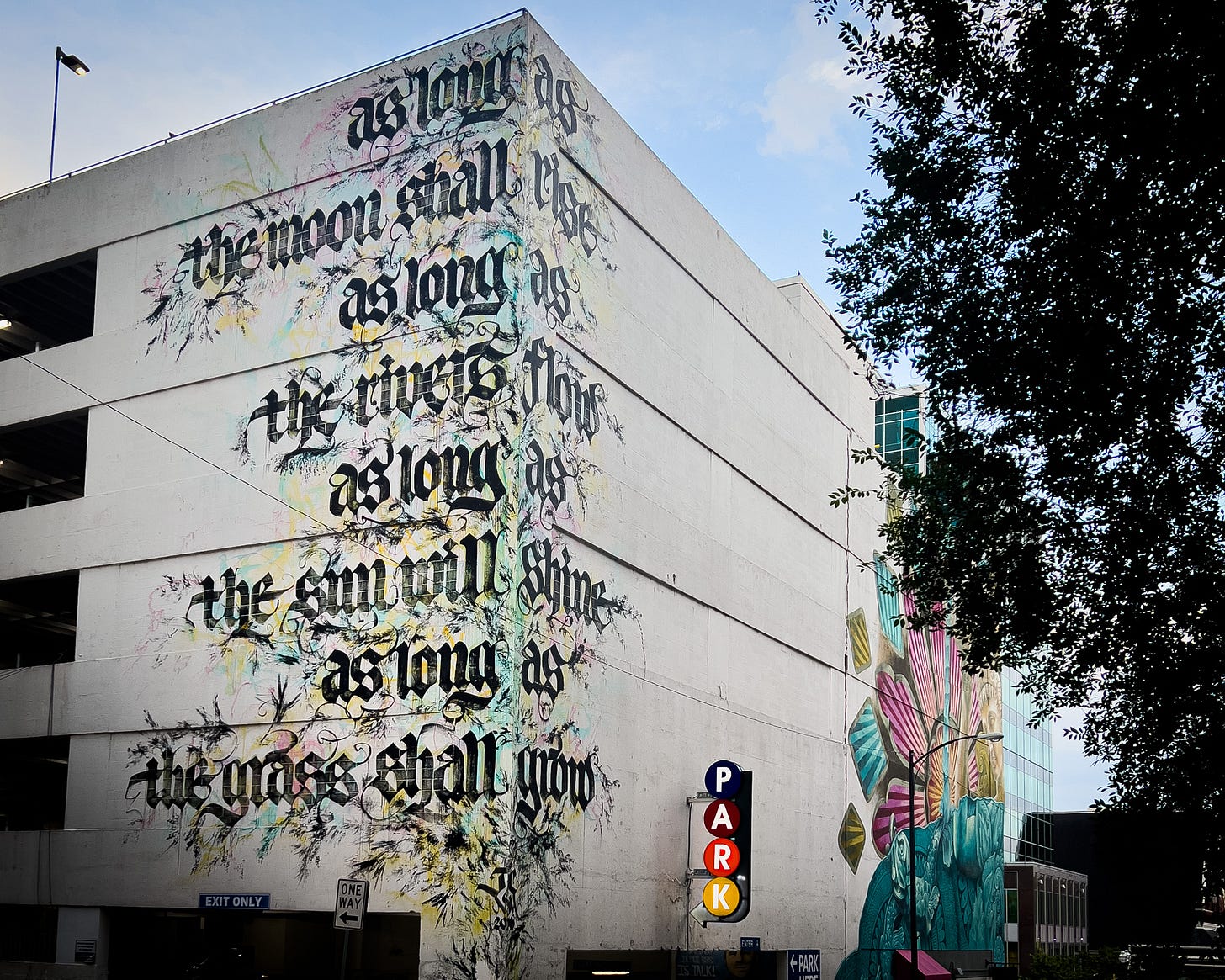 A mural of the the refrain from “As Long As the Grass Shall Grow” on the side of a parking garage in Old English print and swirls: as long as the moon shall rise as long as the rivers flow as long as the sun will shine as long as the grass shall grow