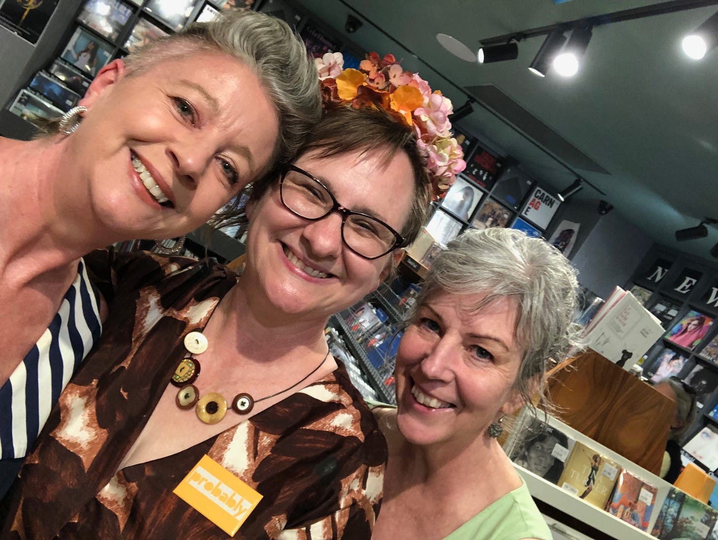 Three smiling women in a bookshop. One of the women is wearing a crown made of hydrangeas. I think she must be some kind of Queen or something. 