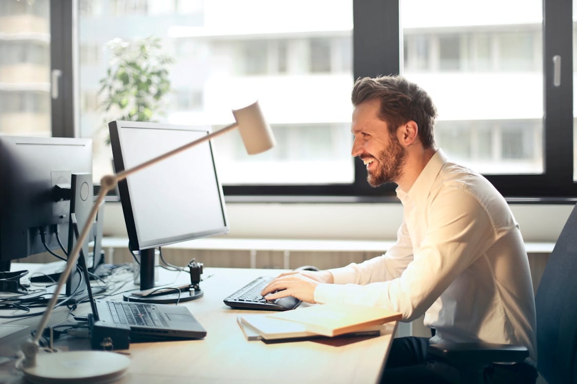 Free Man in White Dress Shirt Sitting on Black Rolling Chair While Facing Black Computer Set and Smiling Stock Photo