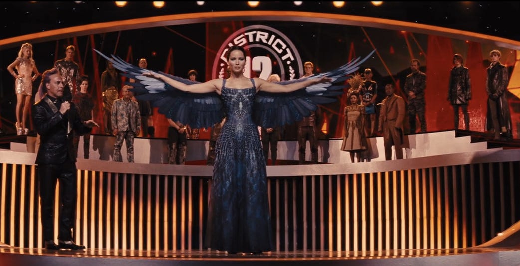 Katniss Everdeen reveals her mockingjay dress onstage, with her arms supporting the wings (Catching Fire, 2013)