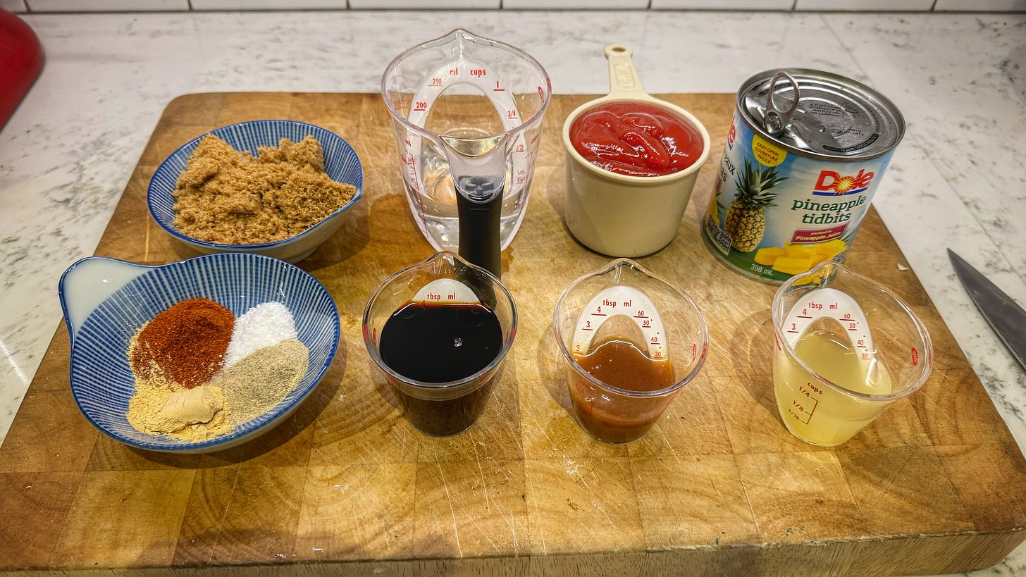 A cutting board with containers of, clockwise from top left: dark brown sugar, white vinegar, ketchup, canned pineapple tidbits, pineapple juice, hot sauce, soy sauce, paprika, kosher salt, white pepper, and garlic powder