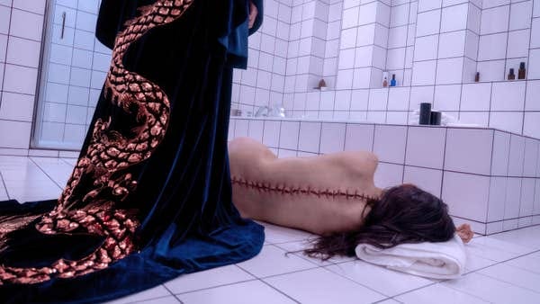 A person with long hair lies face down on a tiled bathroom floor, exposing a stitched incision down their back. Another figure with a snake-patterned garment stands over them