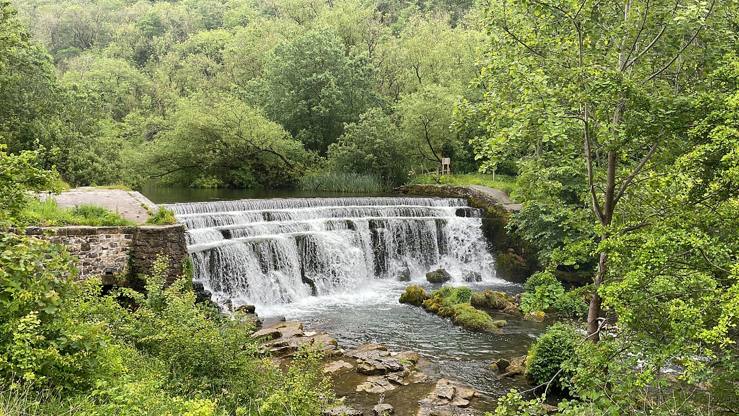 Water flowing over Monsal Dale weir in Derbyshire with leafy trees along the river bank