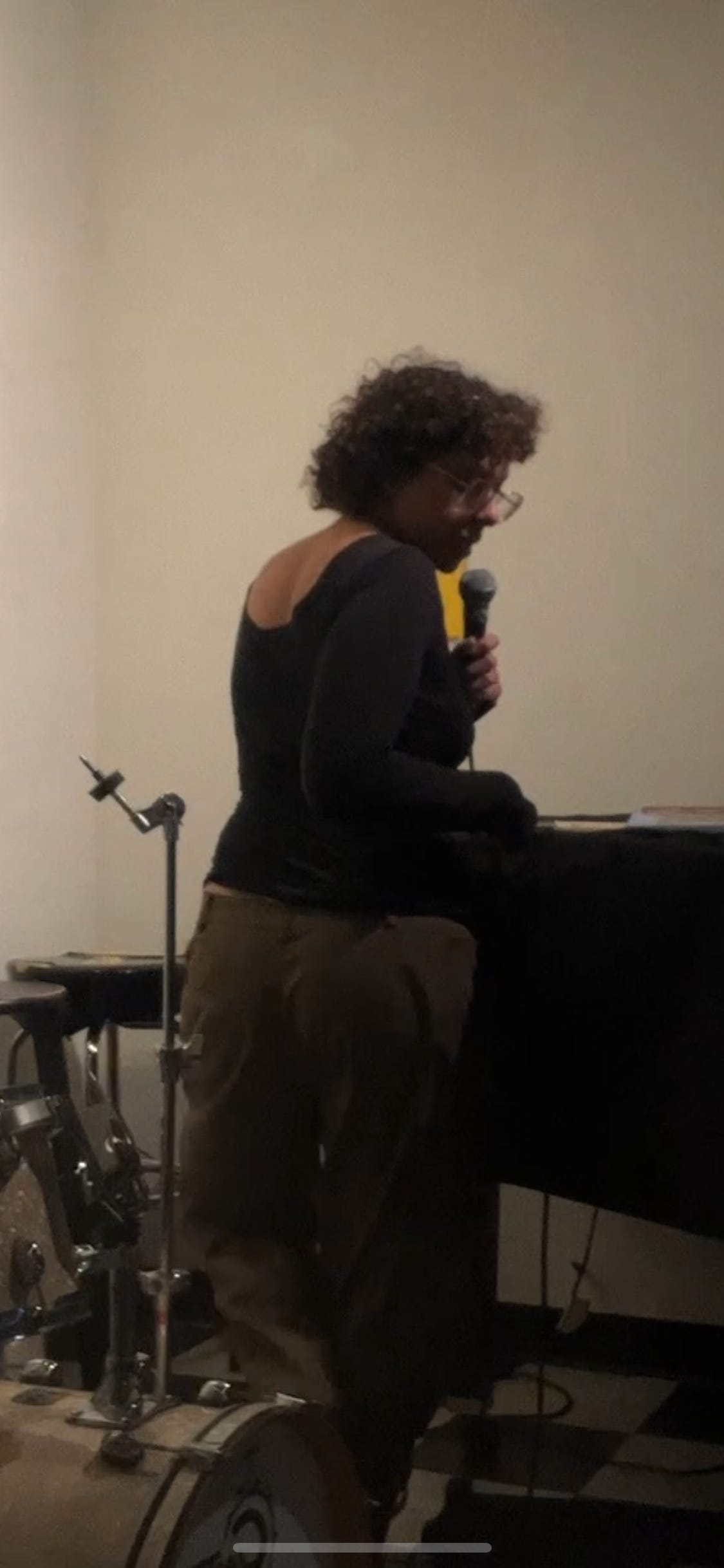 its me! im standing next to a grand piano with my back turned but im turning in a way where you can see i am talking into a microphone. i think i look quite stylish and smart lol