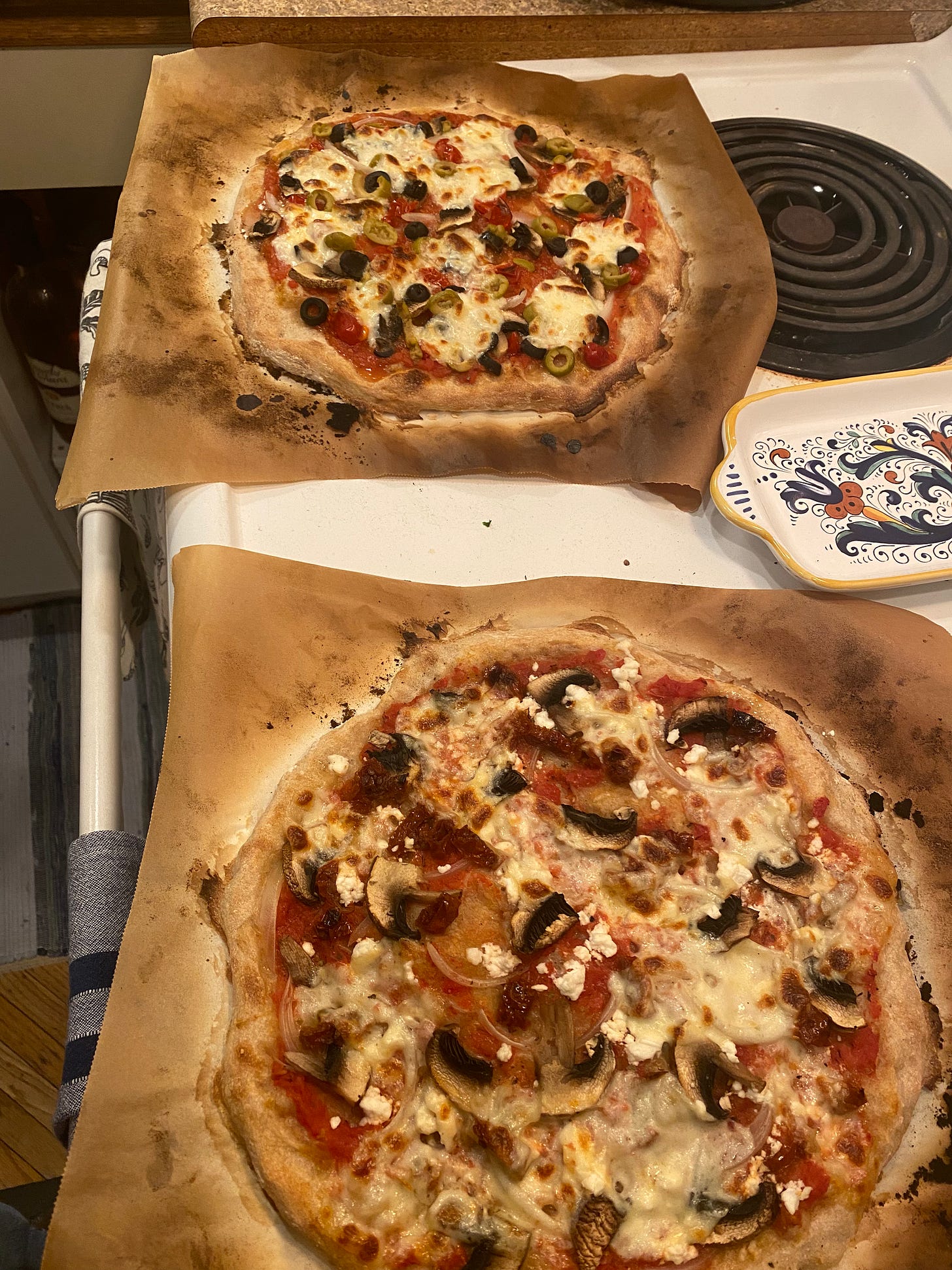 Two pizzas on top of the stove on parchment paper, burned at the edges from the oven. The nearer pizza has mushrooms, feta, and sun-dried tomato, and the one in the background has mushrooms and black & green olive slices.