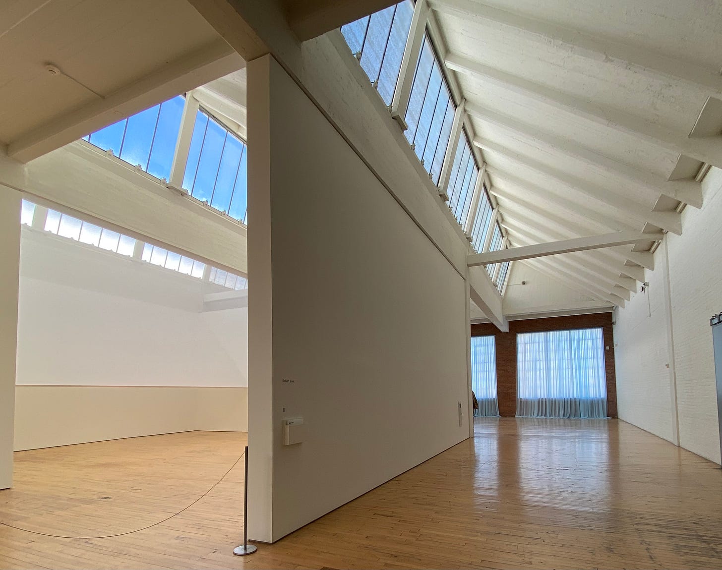 View of Robert Irwin, Full Room Skylight – Scrim V – Dia Beacon, 1972/2022 (right), and Felix Gonzalez-Torres, “Untitled” (Loverboy), 1989 (left), as installed at Dia Beacon. On the left you can see into a gallery room with skylights showing a clear blue sky. The volume of room is subtly divided at an angle from top to bottom by a half-visible pale white scrim. To the right you can see one end of the room where Felix Gonzalez-Torres’s blue curtains have been installed. 