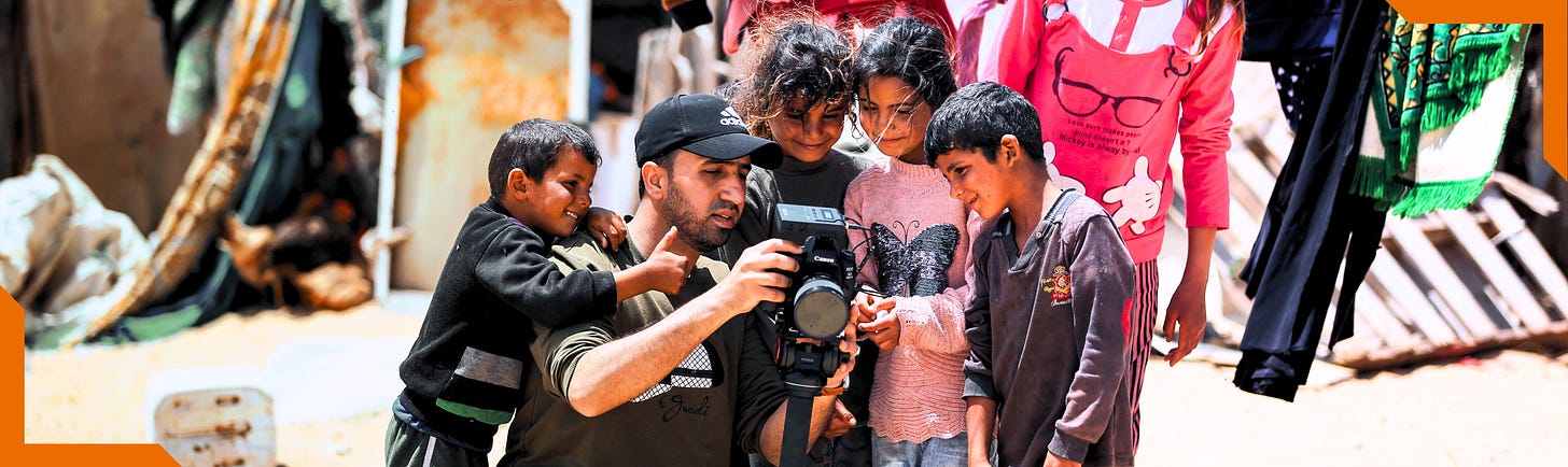 On a sunny day, Palestinian photojournalist Hosny Salah is squatting, holding a camera, in the middle of the frame. Four young children surround him, smiling and looking at the back of his camera. 