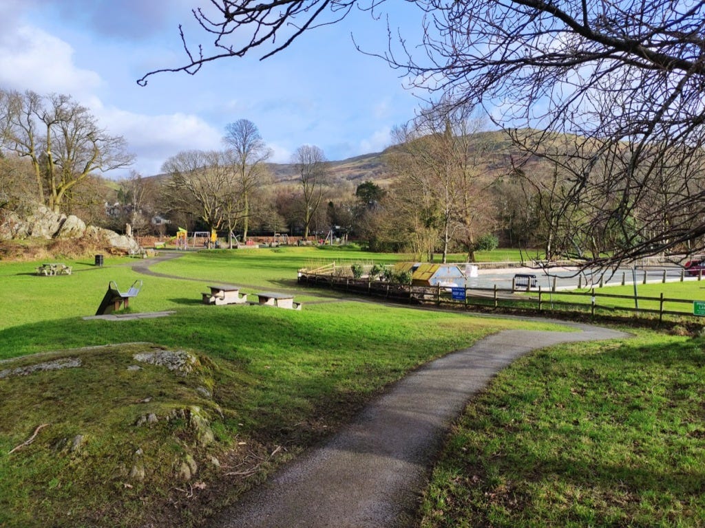 Narrow tarmacced path runs down a small incline, toward a playground in the distance.