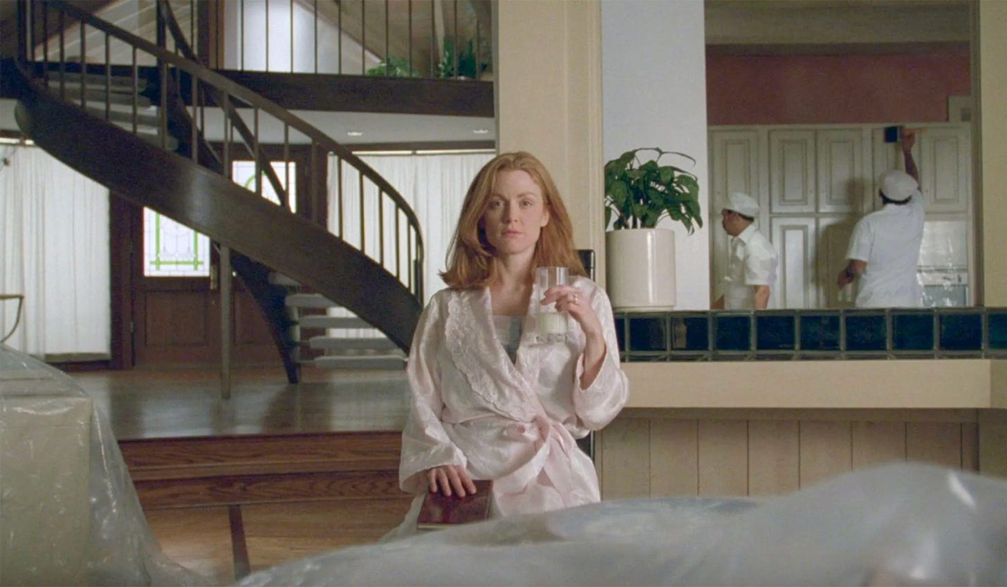 A white woman with long red hair wearing a pink nightgown sits staring into the distance. She has a book on her lap and is holding a glass of milk up with one hand. She’s sat in a large house with a spiral staircase behind, painters renovating and covers still on the furniture.
