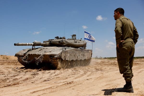 An Israeli soldier in fatigues stands near a tank flying the Israeli flag in the sand.